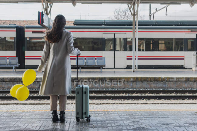 Back view of unrecognizable female traveler in stylish outfit standing with yellow balloons in hand and suitcase on platform of railway station — Stock Photo