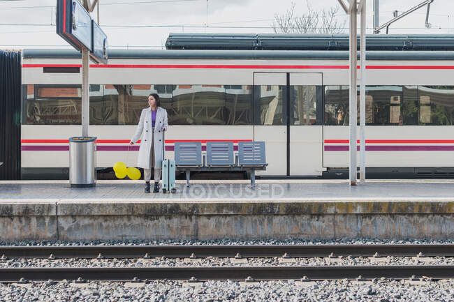 Young female traveler in stylish outfit standing with yellow balloons in hand and suitcase on platform of railway station — Stock Photo