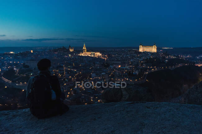 Back view of silhouette of male traveler sitting on hill and enjoying spectacular night cityscape of old city Toledo in Spain with illuminated medieval buildings — Stock Photo