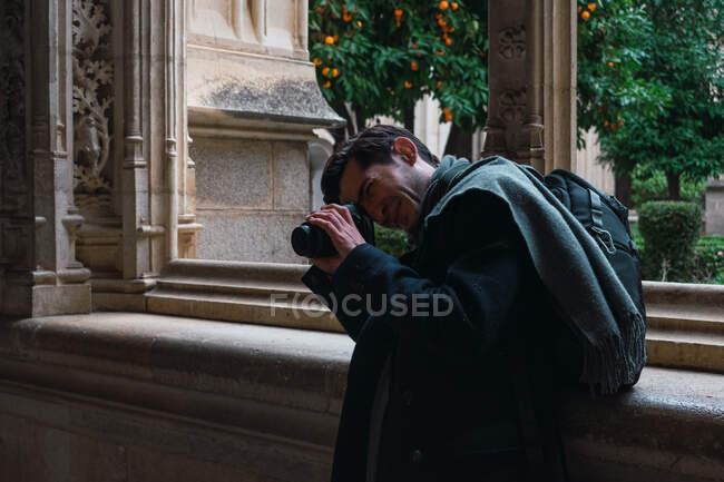 Vista lateral del viajero masculino con la mochila de pie junto a la ventana y tomar una foto con la cámara mientras visita el antiguo edificio de piedra histórica en la ciudad española Toledo - foto de stock