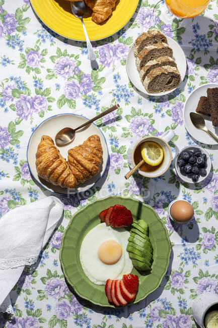 Desayuno casero completo y saludable a la luz del sol con huevos, aguacate, fresas, arándanos, bizcocho, croissants, tostadas, té, café y zumo de naranja - foto de stock