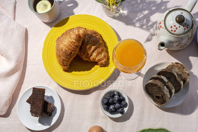 Desde la parte superior de la vista de desayuno completo en el brunch casero a la luz del sol con huevos cocidos, arándanos, bizcocho, croissants, tostadas, té, café y zumo de naranja - foto de stock