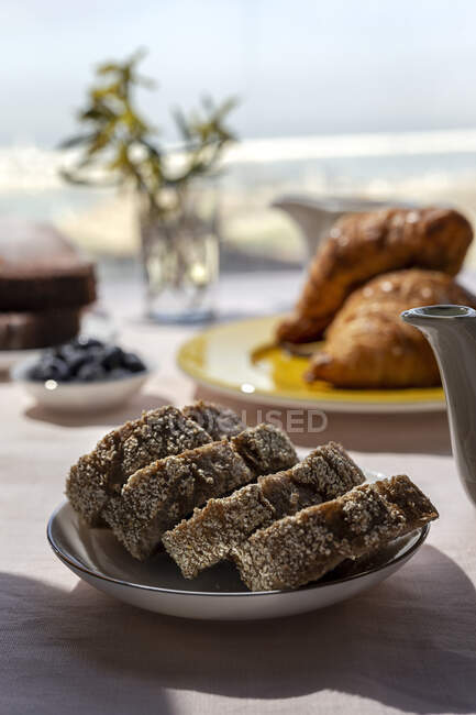 Pequeno-almoço caseiro completo sob luz solar com panela de chá ou café, fatias de pão e croissants — Fotografia de Stock