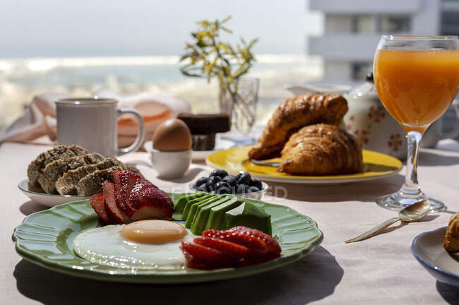 Desayuno casero completo y saludable a la luz del sol con huevos, aguacate, fresas, arándanos, bizcocho, croissants, tostadas, té, café y zumo de naranja - foto de stock