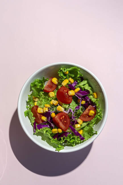 Ensalada de lechuga fresca con tomates de cerezas, cebolla roja y maíz, luz solar sobre fondo rosa desde arriba. Comida saludable. Comida vegana . - foto de stock