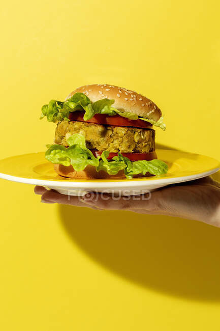 Cropped unrecognizable person hand holding a homemade vegan green lentil burger with tomato, lettuce and french fries on a yellow colorful background — Stock Photo