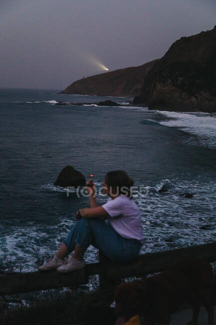 From above side view of casual pensive young female smoking cigarette while sitting on fence on rocky sea coast in summer evening in town of Lekeitio in Spain with beacon light in background — Stock Photo