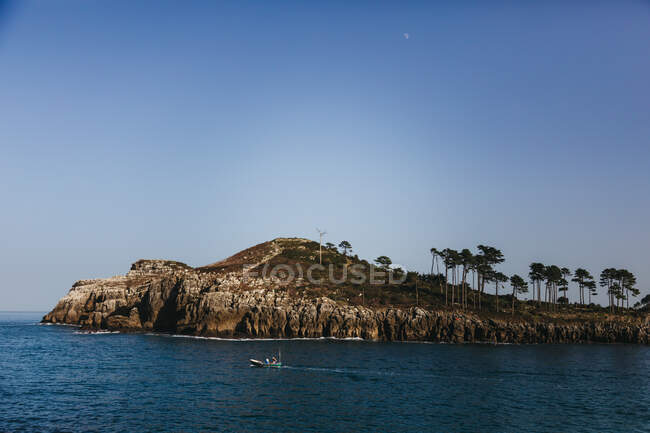 Barco de pesca solitário movendo-se na baía com água tranquila contra a costa rochosa com árvores verdes altas a pé da colina sob céu azul claro em Espanha — Fotografia de Stock