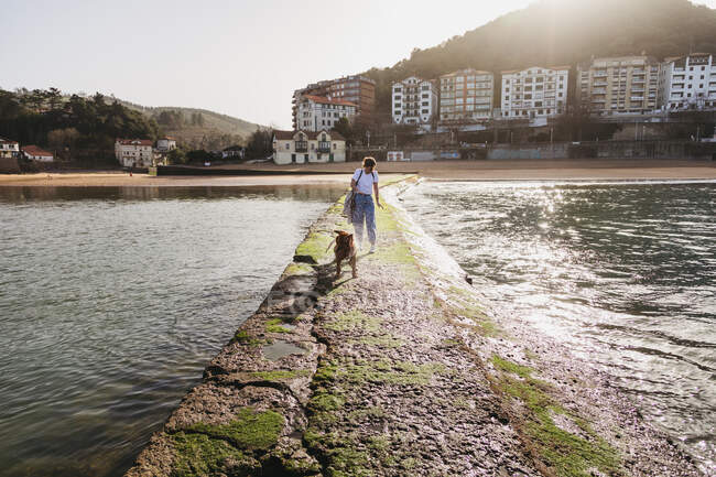 Jeune femme promenant avec chien sur une jetée en pierre dans la ville de Lekeitio au Pays Basque avec des bâtiments résidentiels et des collines en arrière-plan — Photo de stock