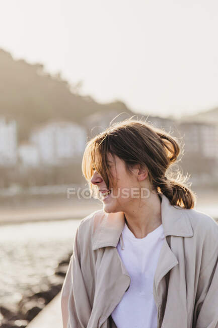 Happy blond female in casual clothes looking away laughing while standing on pier and touching hair against blurred urban environment of resort town in Spain — Stock Photo
