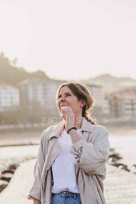Glückliche blonde Frau in lässiger Kleidung, die lachend wegschaut, während sie auf der Seebrücke steht und die Haare vor der verschwommenen städtischen Umgebung des Ferienortes in Spanien berührt — Stockfoto