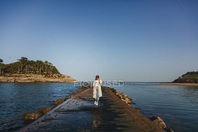 Vista trasera de una viajera sin rostro vestida con ropa casual caminando por el muelle de madera mojada contra las olas del mar y la costa rocosa en España - foto de stock
