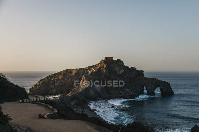Empty road leading along bridge and ridge to lonely country house on top of rocky hill on island Gaztelugatxe surrounded by calm sea water with white foam waves in sunny evening — Stock Photo