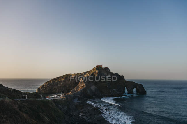 Paving stone way leading along stone bridge and ridge of rocky hill to lonely country house on island Gaztelugatxe surrounded by tranquil sea water with white foam waves — Stock Photo