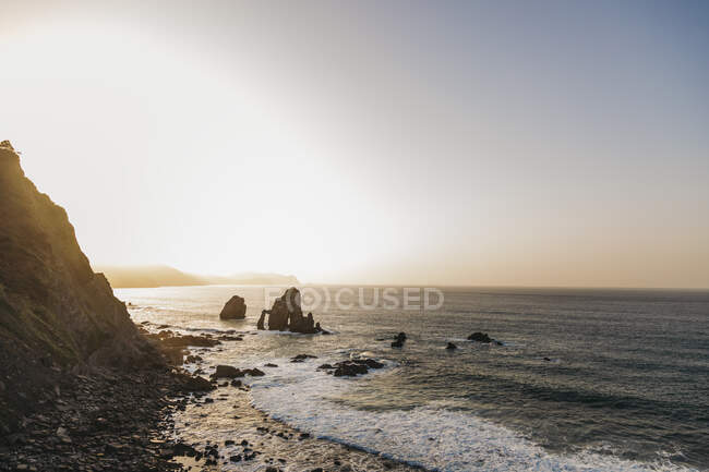 Schöne Meereslandschaft mit klarem blauem Wasser und unruhigen weißen Schaumwellen am Strand gegen bewaldete Hügel an der Küste und einsame Felseninsel unter wolkenlosem Himmel in Spanien bei Sonnenuntergang — Stockfoto