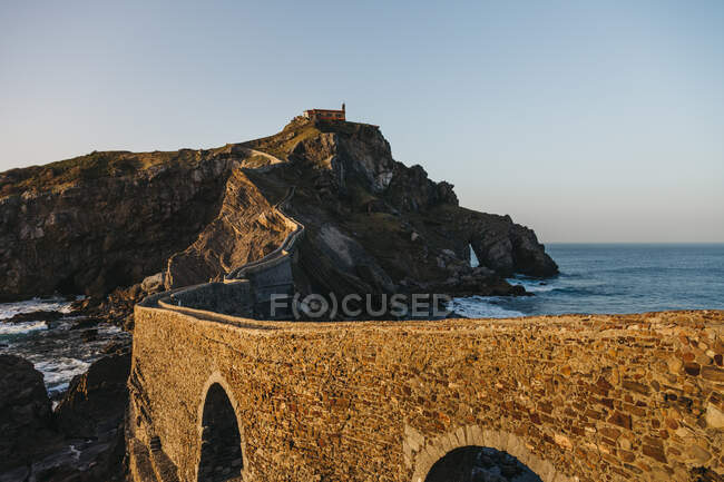 Empty road leading along stone bridge and ridge of rocky hill to lonely country house on island Gaztelugatxe surrounded by tranquil sea water with white foam waves during sunset — Stock Photo