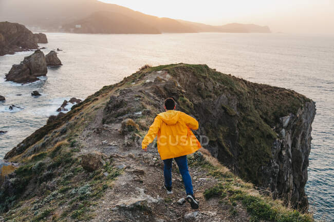 Back view of unrecognizable man in vibrant yellow jacket and denim running on rocky hill and enjoying picturesque scenery of sea coast during sunset in Spain — Stock Photo