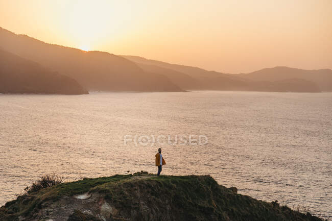 Hombre vista lateral en chaqueta amarilla vibrante y denim de pie en la colina rocosa y disfrutar de pintorescos paisajes de la costa del mar durante el atardecer en España - foto de stock
