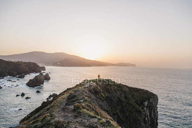 Visão traseira do homem irreconhecível com jaqueta amarela vibrante e ganga em pé na colina rochosa e desfrutando de paisagem pitoresca da costa do mar durante o pôr do sol na Espanha — Fotografia de Stock