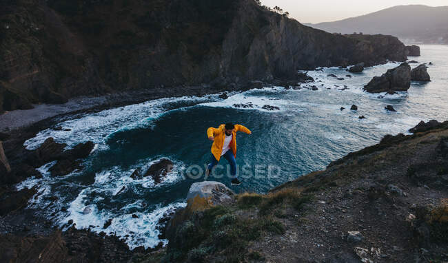De cima pessoa na jaqueta amarela vibrante pulando na borda do penhasco e desfrutando de paisagem incrível da costa do mar rochoso durante o pôr do sol na Espanha — Fotografia de Stock