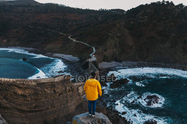 From above back view of faceless man in vivid yellow jacket and denim standing on edge of cliff and looking at empty road crossing old stone bridge among boulders and troubled waves of bay to forested hills in Spain — Stock Photo
