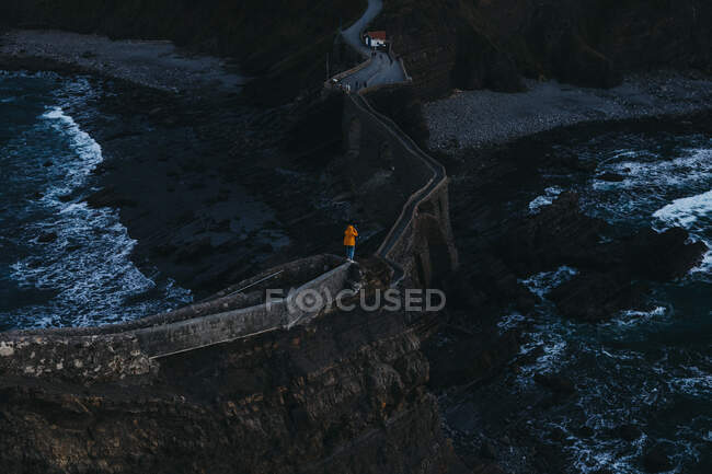 Von oben Rückenansicht einer Person mit ausgestreckten Armen, die die Freiheit genießt, auf einer alten Steinbrücke stehend gegen aufgewühltes Wasser mit Schaumwellen, die felsiges Ufer in Spanien spülen — Stockfoto