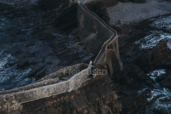 Dall'alto donna irriconoscibile con animale domestico in piedi sul vecchio ponte di pietra contro l'acqua travagliata baia lavaggio costa rocciosa in Spagna — Foto stock