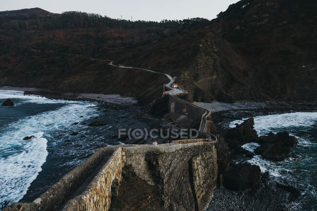 Извилистая горная дорога, пересекающая каменный мост и ведущая вдоль скалистого побережья Испании в сумерках — стоковое фото