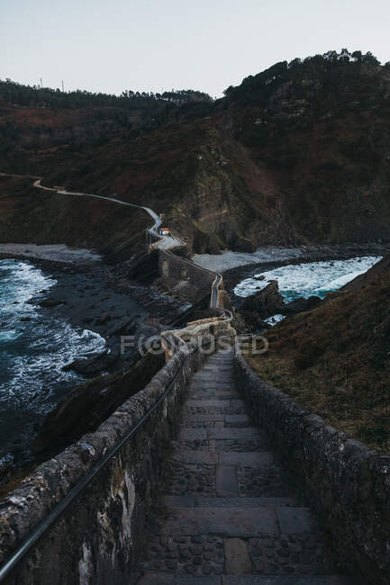 Пустой тротуарный камень, ведущий вдоль моста среди каменных заборов и склонов скалистого холма вдоль берега, омываемый морской водой с белой пеной в сумерках в Испании — стоковое фото