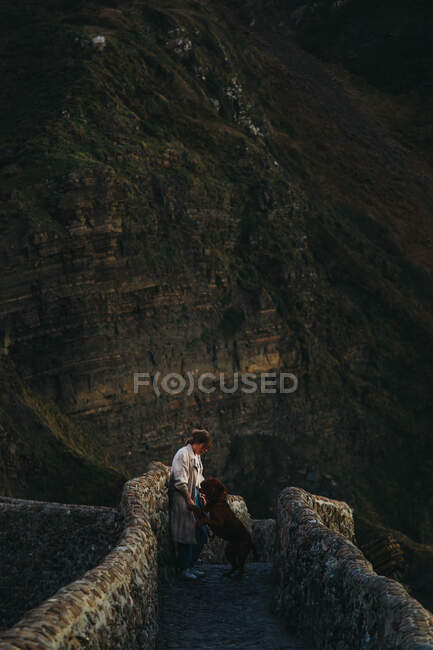 Высокоугольный вид сбоку женщины в повседневной одежде с большой коричневой собакой, стоящей на старом каменном мосту и смотрящей друг на друга на каменистом склоне горы в Испании — стоковое фото