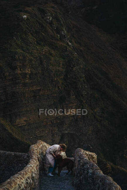 Висококутний вид на самицю в повсякденному одязі з великим коричневим собакою стоїть на старому кам'яному мосту і дивиться один на одного проти скелястого схилу гори в Іспанії. — стокове фото