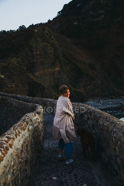 Rückansicht einer Frau in Freizeitkleidung mit einem großen braunen Hund, die auf einer alten Steinbrücke steht und auf einen felsigen Berghang in Spanien blickt — Stockfoto