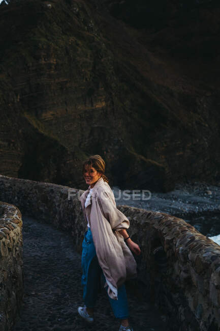 Femme en vêtements décontractés marchant sur un vieux pont de pierre souriant et regardant la caméra contre l'eau de baie troublée laver la côte rocheuse en Espagne — Photo de stock
