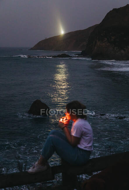 From above side view of casual pensive young female smoking cigarette while sitting on fence on rocky sea coast in summer evening in town of Lekeitio in Spain with beacon light in background — Stock Photo