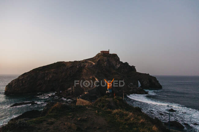 Cheerful person jumping against road crossing old stone bridge and leading to lonely country house on rocky island Gaztelugatxe during sunset in Spain — Stock Photo