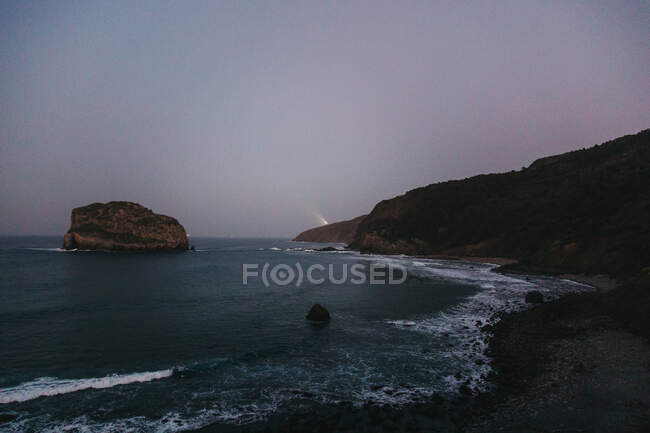 Hermoso paisaje marino con agua azul clara y olas de espuma blanca en la playa contra colinas boscosas en la costa y la isla rocosa solitaria bajo el cielo despejado en España durante la puesta del sol - foto de stock