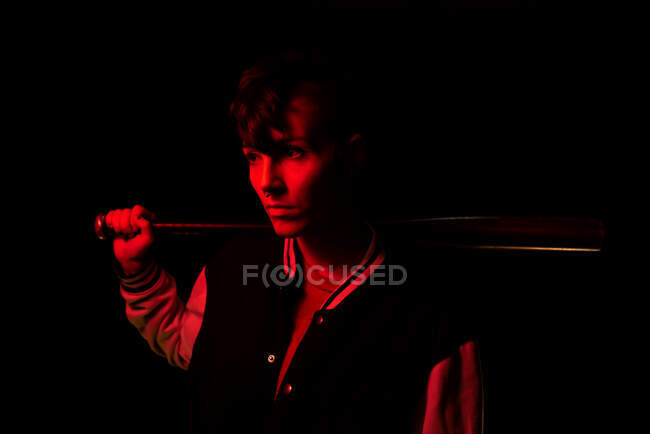 Side view of contemporary female in bomber jacket looking away holding a black baseball bat with red light on background — Stock Photo