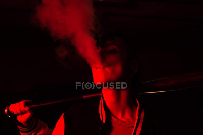 Zeitgenössische Frau in Bomberjacke mit schwarzem Baseballschläger auf der Schulter beim Rauchen mit rotem Licht im Hintergrund — Stockfoto