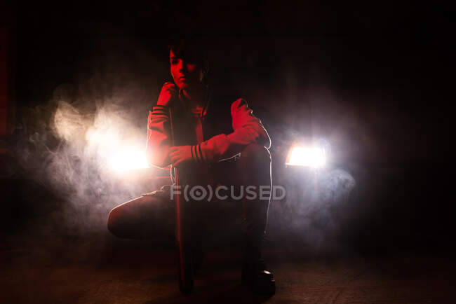 Young hipster woman in jeans and black boots with massive sole looking away while holding bat with car headlights on background — Stock Photo