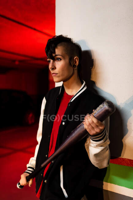 Contemporary female in bomber jacket looking away while leaning on wall with black baseball bat on shoulder with red light on background — Stock Photo