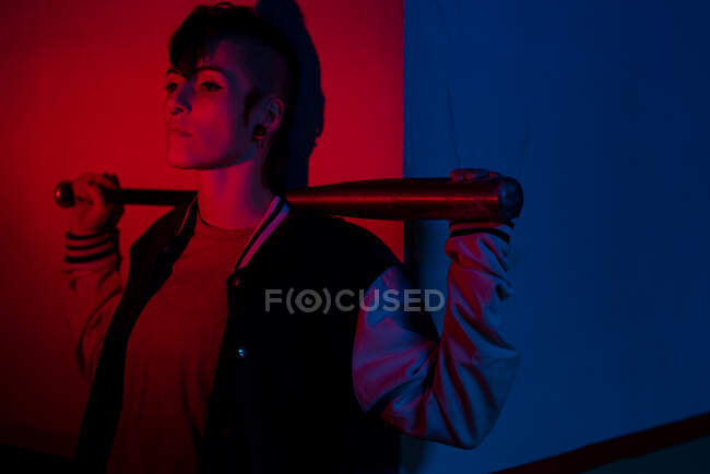 Mujer poco convencional con peinado moderno de pie contra la pared con bate de béisbol detrás de la cabeza y mirando hacia otro lado iluminado por rojo y azul en la calle de la ciudad - foto de stock