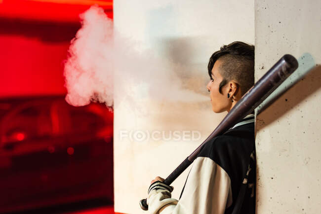 Side view of contemporary female in bomber jacket leaning on wall with black baseball bat on shoulder while smoking with red light on background — Stock Photo