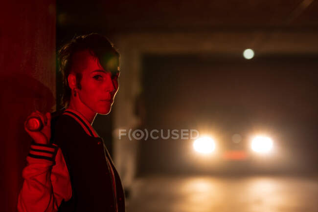 Side view of contemporary female in bomber jacket holding a black baseball bat on shoulder looking at camera with red light and car lights on background — Stock Photo