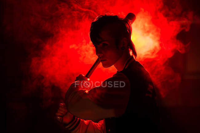 Femme rebelle en veste décontractée avec piercing et coiffure moderne tenant chauve-souris parmi la lumière rouge colorée et la vapeur — Photo de stock