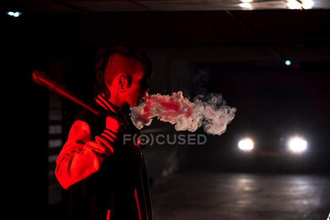 Side view of contemporary female in bomber jacket holding a black baseball bat on shoulder while smoking with red light and car lights on background — Stock Photo