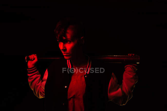 Vista laterale della donna contemporanea in giacca bomber guardando lontano con una mazza da baseball nera con luce rossa sullo sfondo — Foto stock