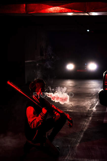 Vue latérale d'une femme contemporaine méconnaissable accroupie dans une veste de bombardier tenant une batte de baseball noire sur l'épaule tout en fumant avec une lumière rouge et des feux de voiture sur le fond — Photo de stock