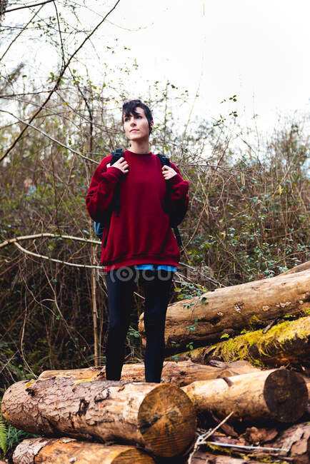Woman in casual clothes and sneakers with backpack while standing on wooden logs and resting on hill slope among greenery in forest in daytime in Spain — Stock Photo