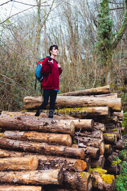 Donna in abiti casual e scarpe da ginnastica con zaino mentre in piedi su tronchi di legno e poggiata su pendio collinare tra il verde nella foresta durante il giorno in Spagna — Foto stock