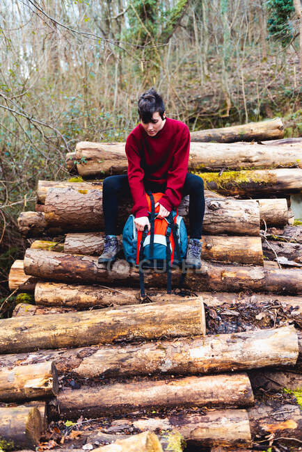 Low angle of woman in casual clothes and sneakers opening backpack while sitting on wooden logs and resting on hill slope among greenery in forest in daytime in Spain — Stock Photo